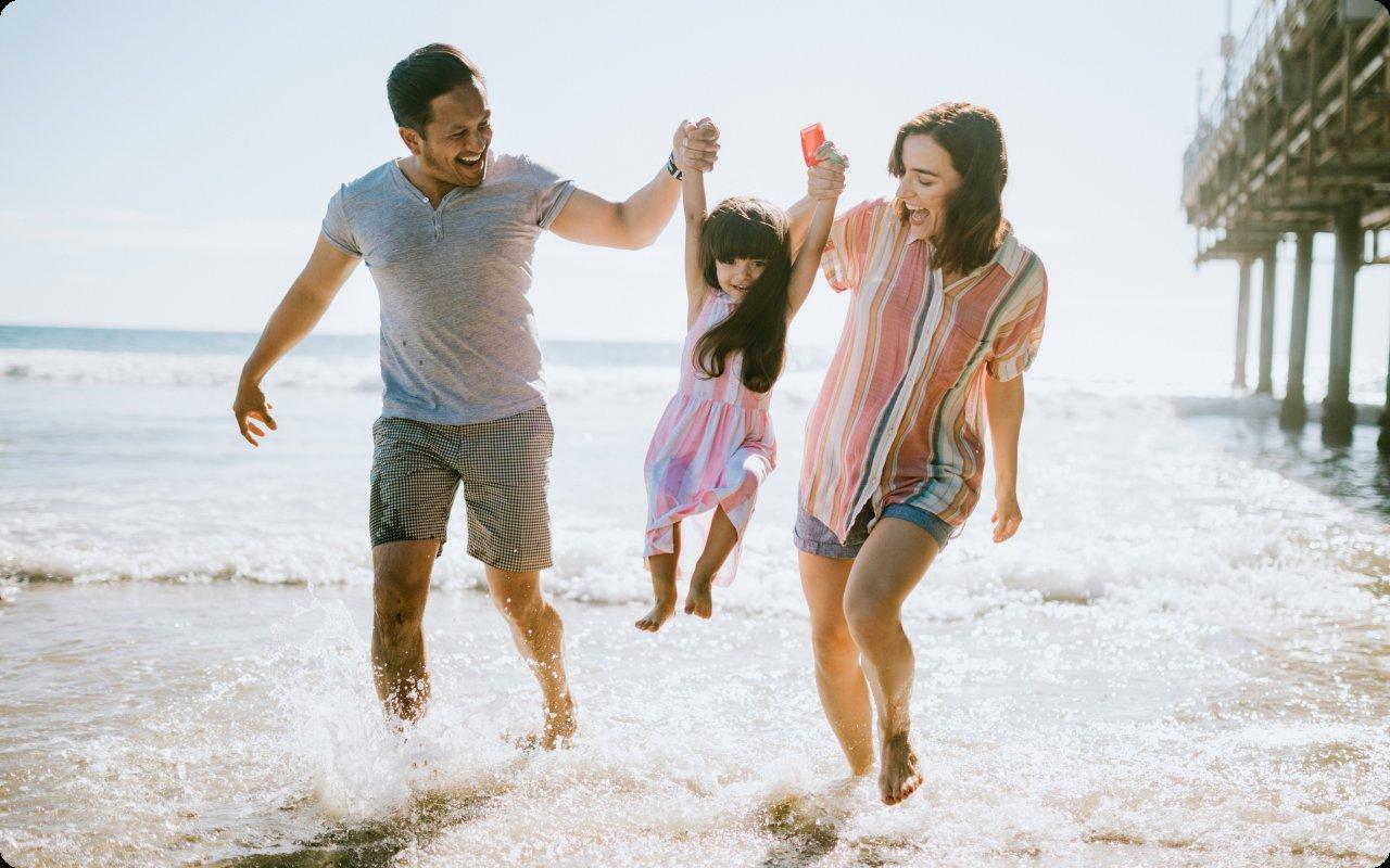 Parents swinging their daughter by the hands in the surf beside a beach pier