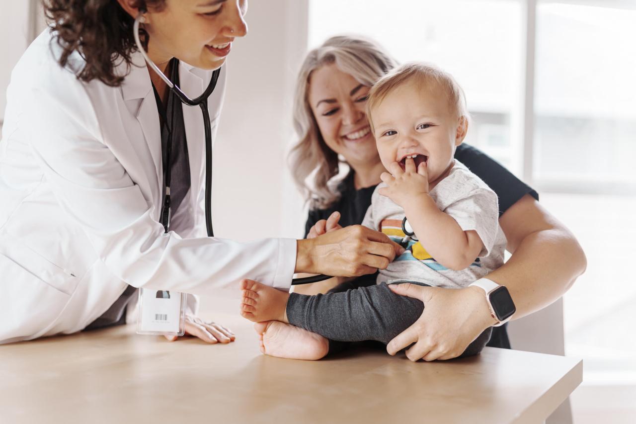 A pediatrician listens to a smiling baby's chest with a stethoscope as the baby's mother holds him