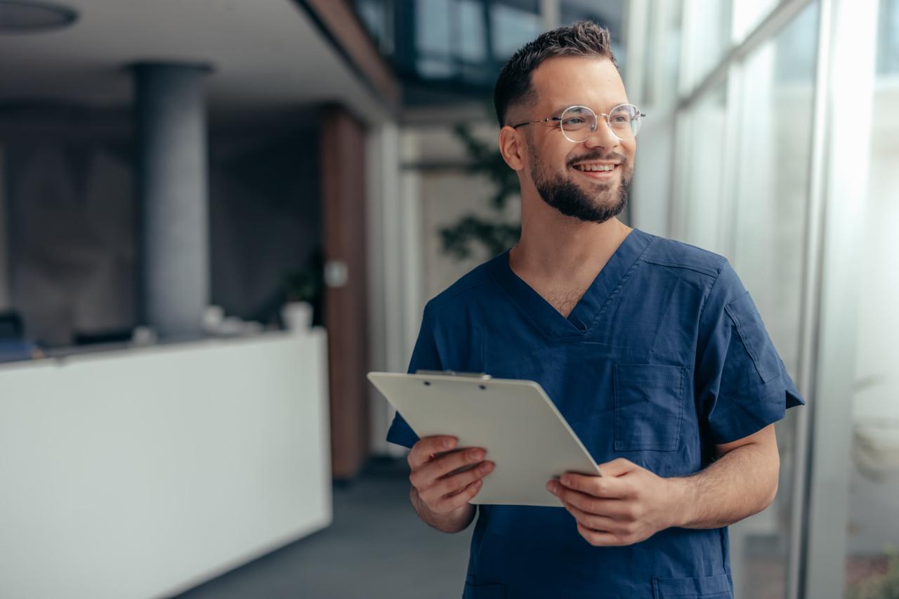 A smiling clinical staff member in scrubs holds a clipboard in a practice hallway